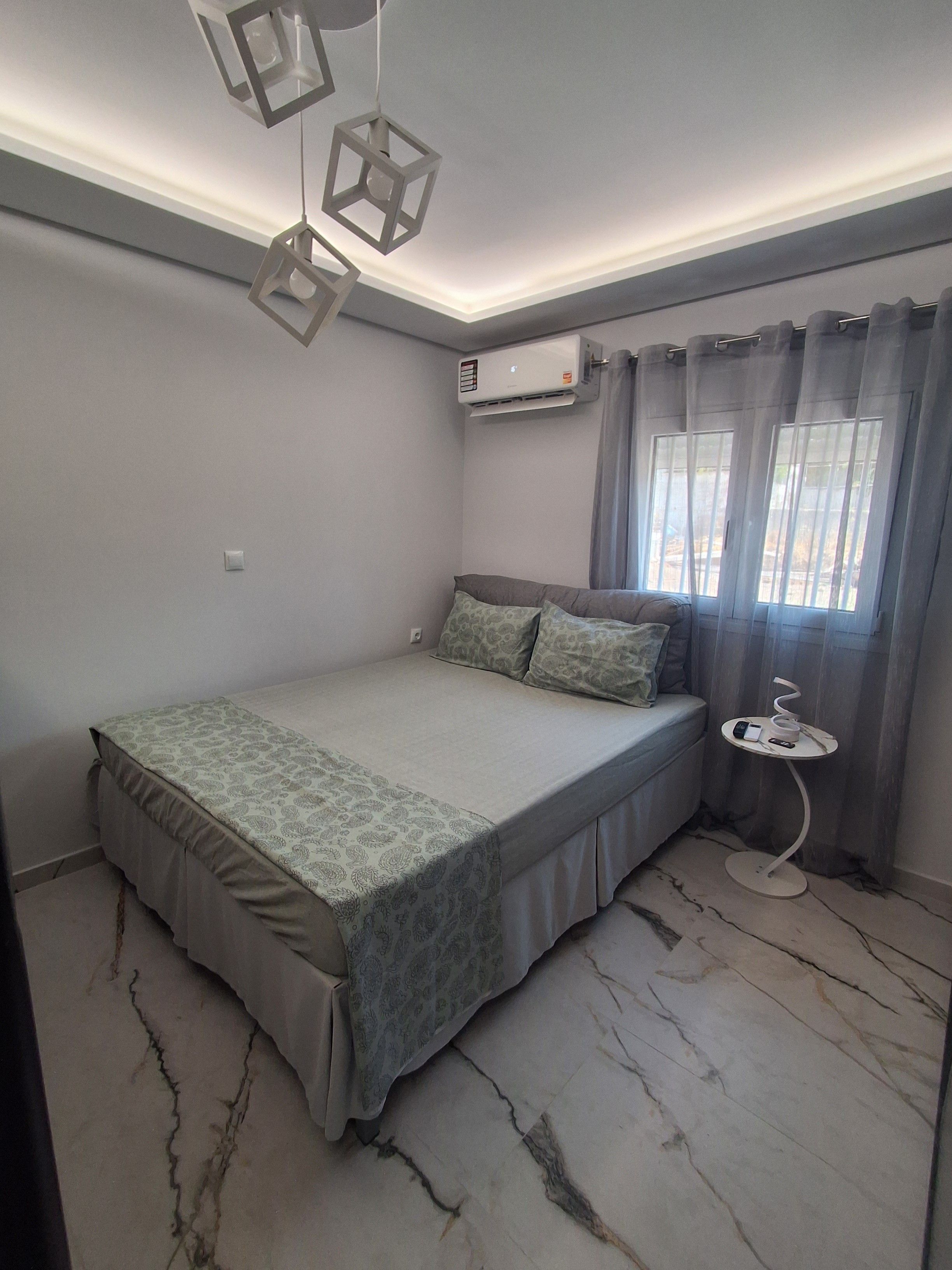 Une chambre à coucher avec un lit, une table de chevet sur laquelle se trouvent un téléphone et une tasse, une fenêtre avec des rideaux et un appareil de climatisation.