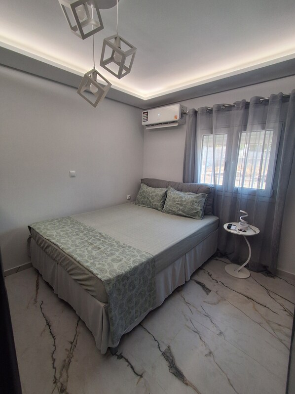 Une chambre à coucher avec un lit, une table de chevet sur laquelle se trouvent un téléphone et une tasse, une fenêtre avec des rideaux et un appareil de climatisation.