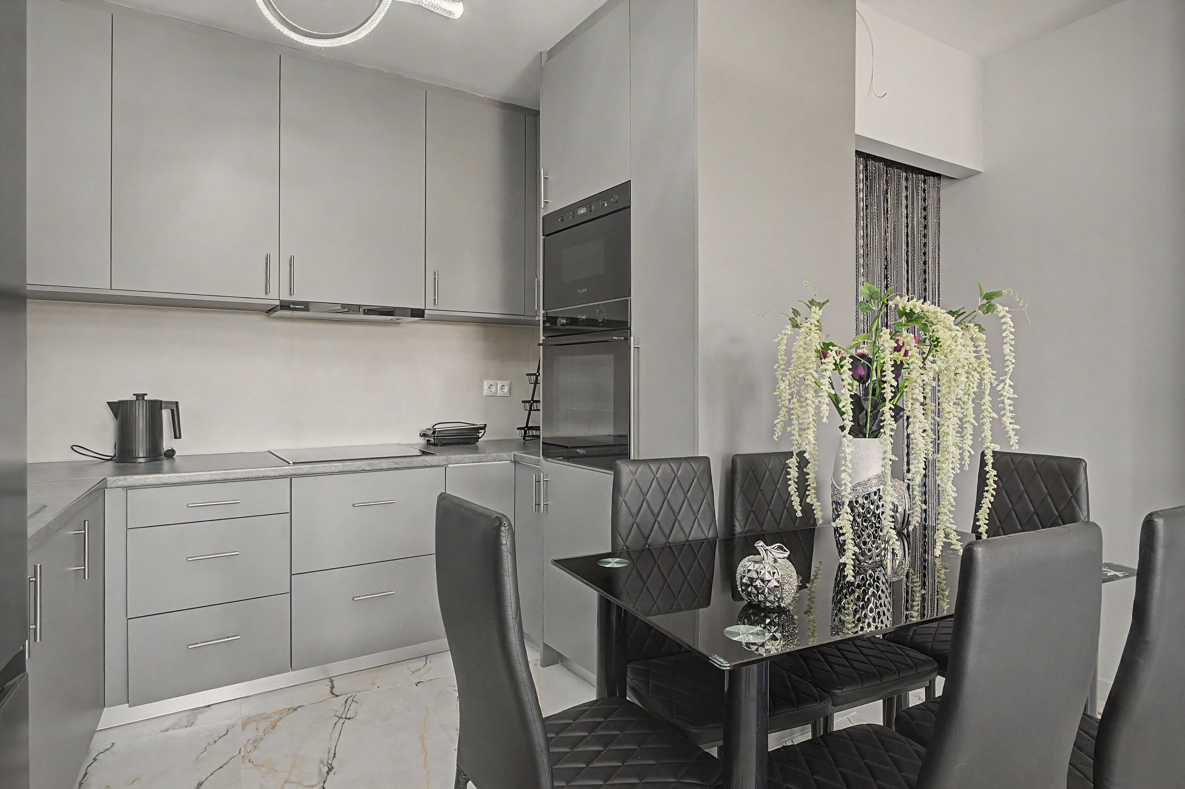 Une cuisine moderne avec des meubles gris, un four encastré et un coin repas comprenant une table et des chaises noires.