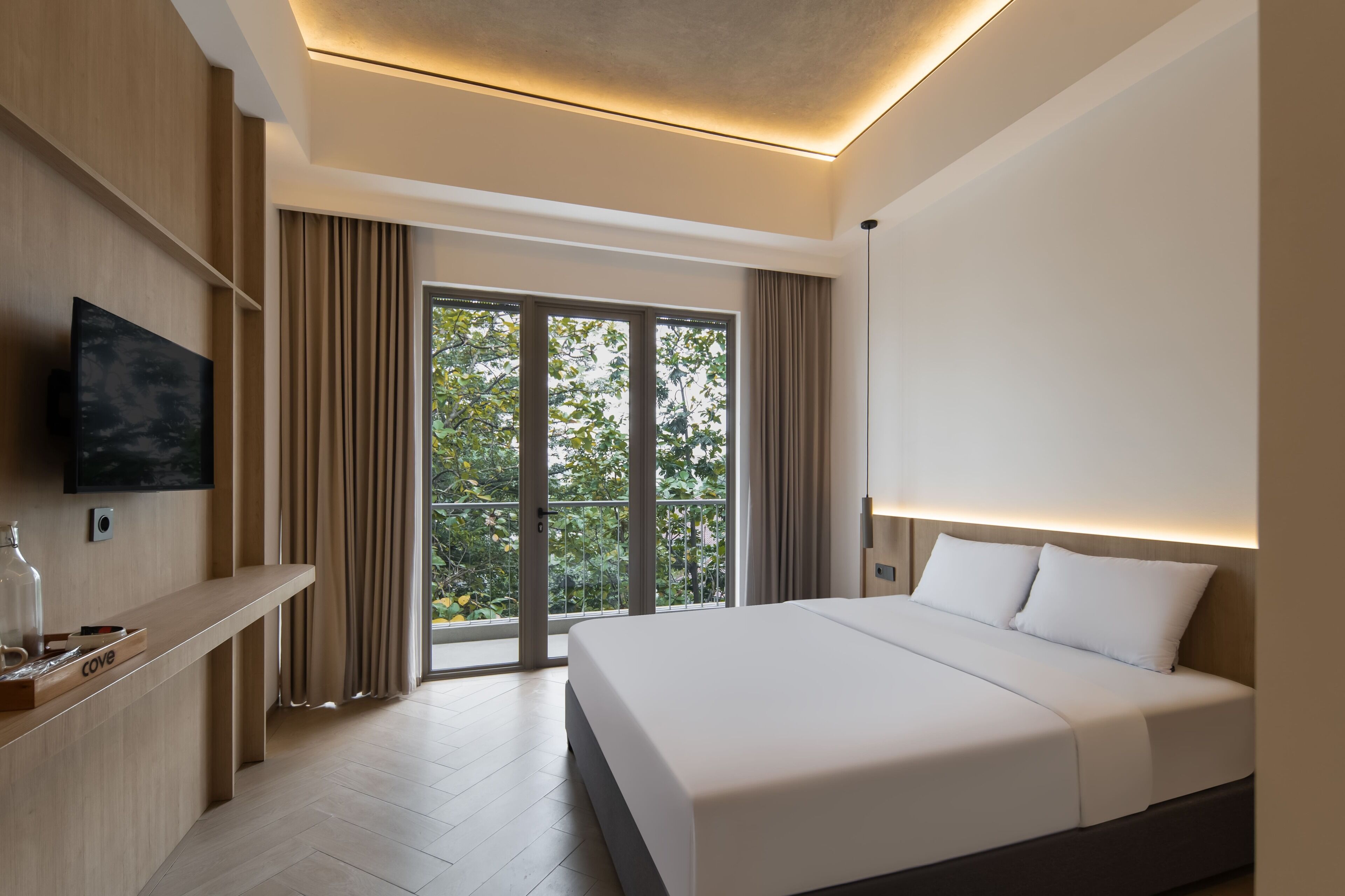 A modern hotel room with a large bed, a flat-screen TV mounted on the wall, and a balcony with a view of trees.