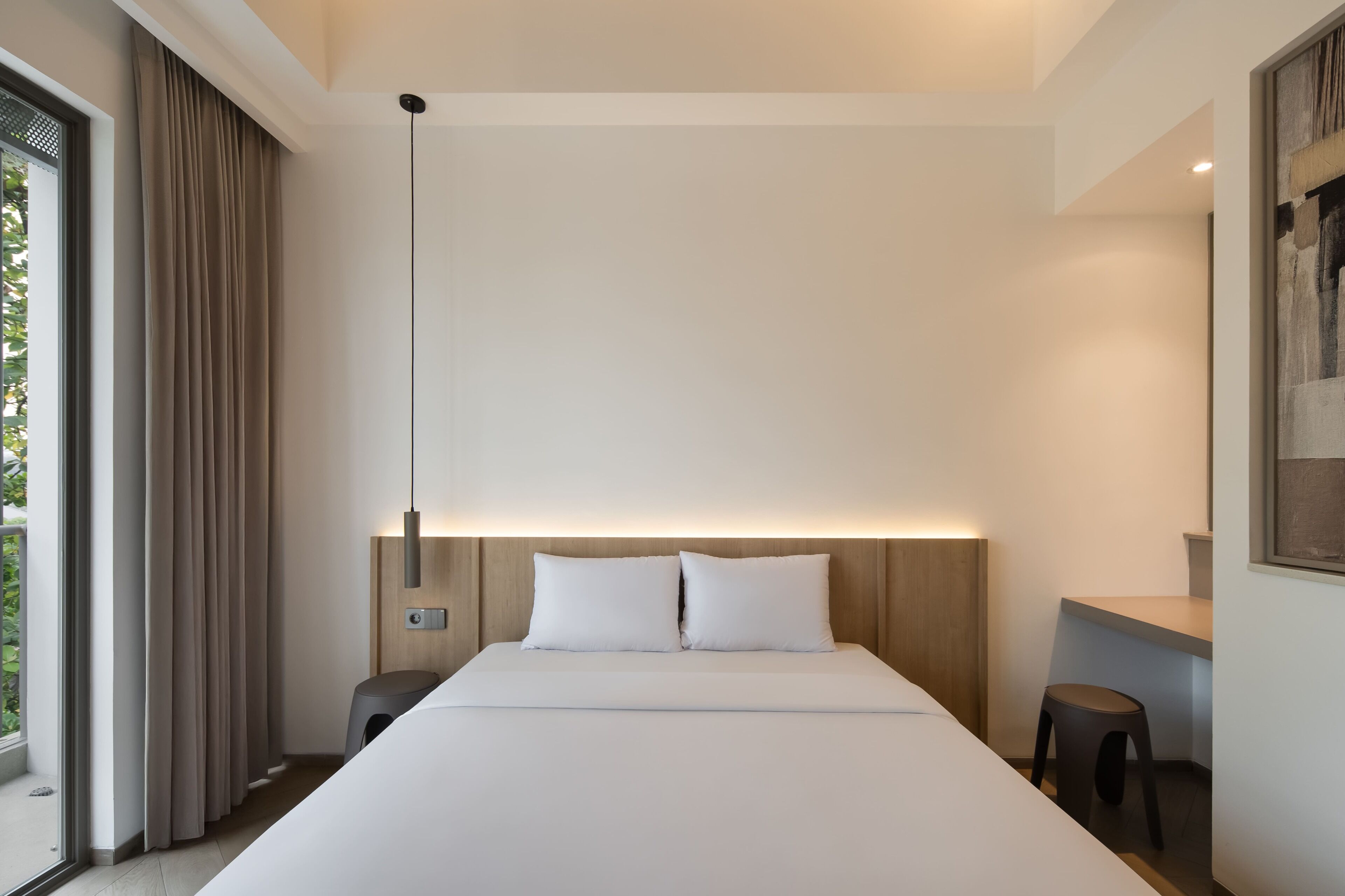 A modern hotel room with a large bed, a bedside table, a nightstand, a window with curtains, and a wall-mounted light fixture.