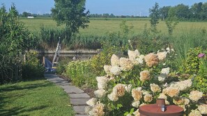 Een tuinpad dat leidt naar een rustieke buitenruimte met een metalen constructie, omringd door weelderige begroeiing en bloeiende planten.