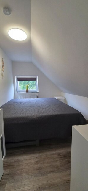 Een klein appartement met één kamer, schuine plafonds, een bed, een radiator en een raam met uitzicht op groen.