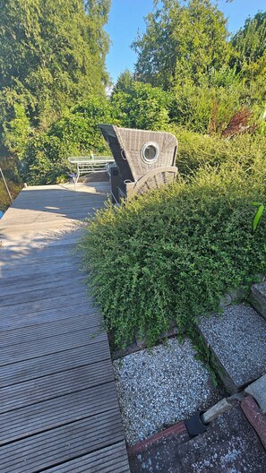 A wooden deck with a wicker chair and a table, surrounded by greenery and a pebble path. - Relax by the Water – Vacation Home with Its Own Boat Dock in Emden (Emden)