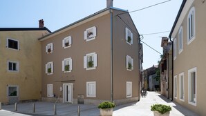 Uma rua estreita com edifícios que apresentam várias janelas e persianas, uma porta e defensas.