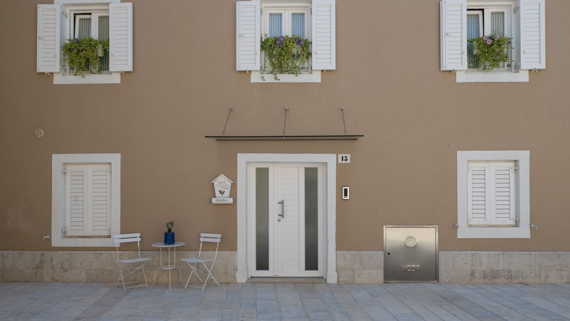 A building facade with a central door, numbered 15, flanked by windows with shutters and flower boxes.