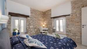 Quarto com parede de pedra, cama com colcha estampada azul, escrivaninha com cadeira e janela com cortinas.