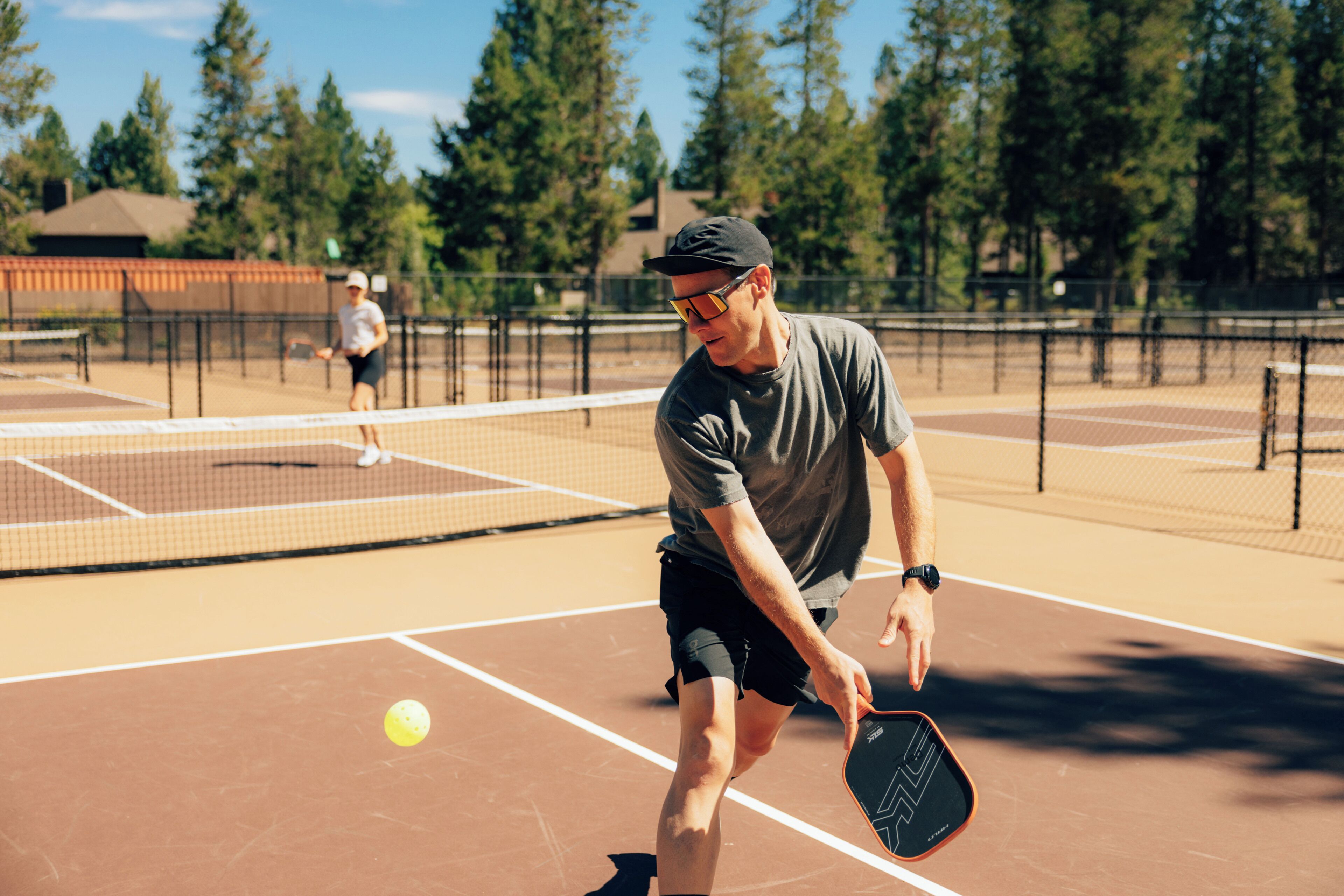 A tennis court with a net, a player in a cap and sunglasses, and another player in the background.