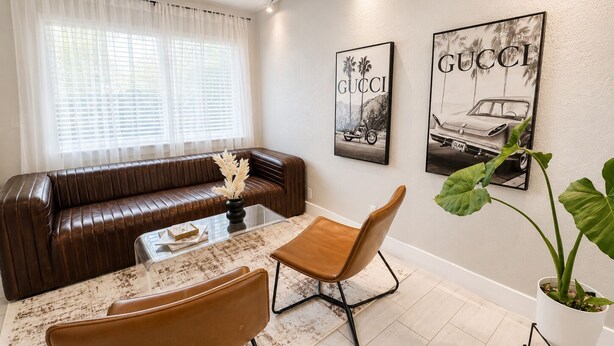Un salón con un sofá de cuero marrón, una mesa de centro de vidrio, dos sillones de cuero marrón y dos carteles de Gucci colgados en la pared.