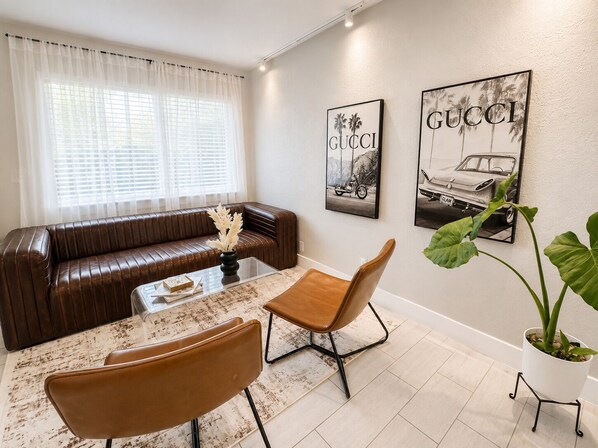 A living room with a brown leather sofa, a glass coffee table, two brown leather chairs, and two wall-mounted Gucci posters.