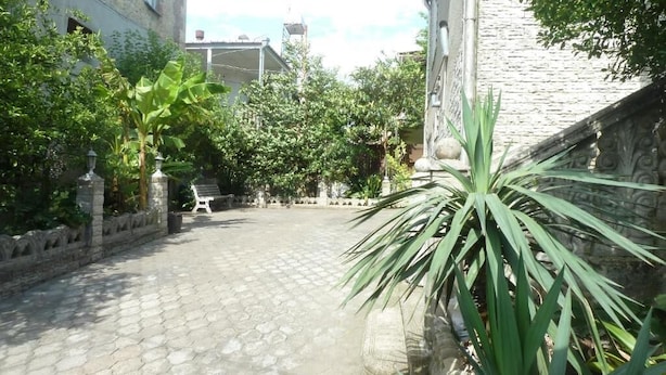 Un patio con pavimento de adoquines, rodeado de muros de piedra y abundante vegetación, entre la que se observan plataneros y una palmera.