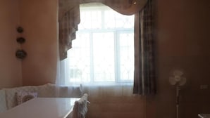 Una habitación con cama, una mesa, una ventana con cortinas y un ventilador de pie.