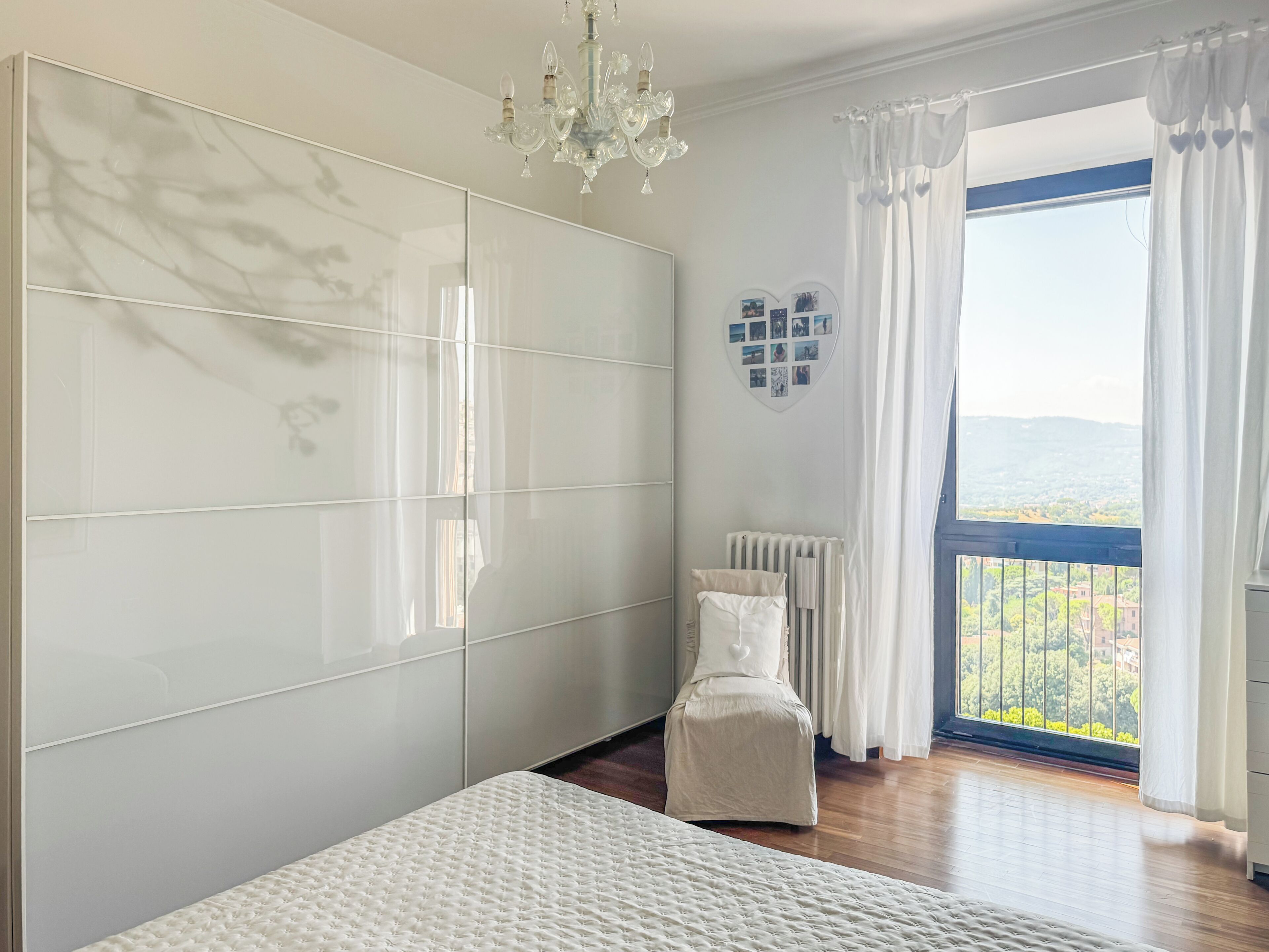 A bedroom with a large bed, a chest of drawers, a radiator, and a view of the outdoors.