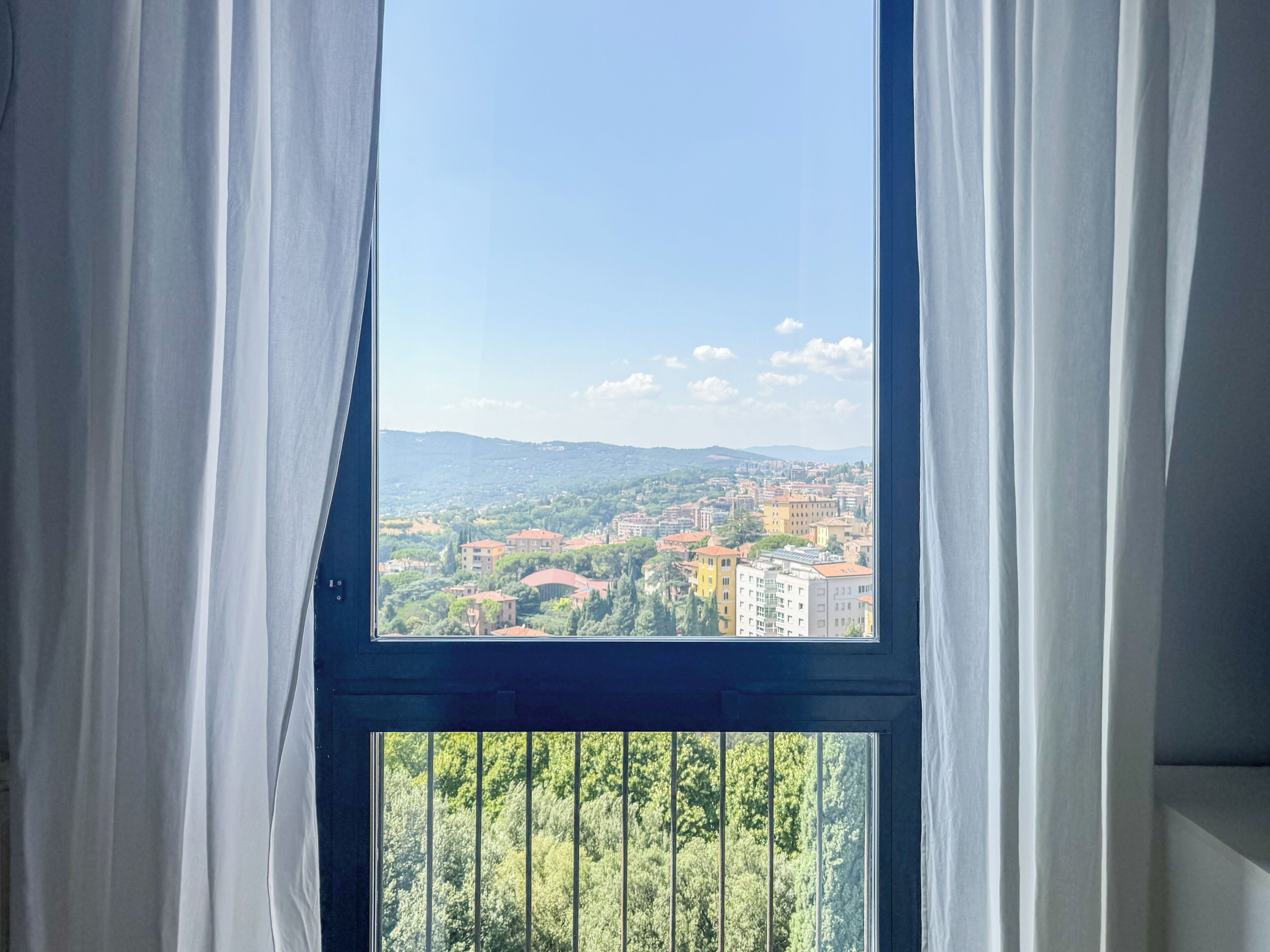 A window with a view of a cityscape, curtains partially drawn, and a balcony railing.