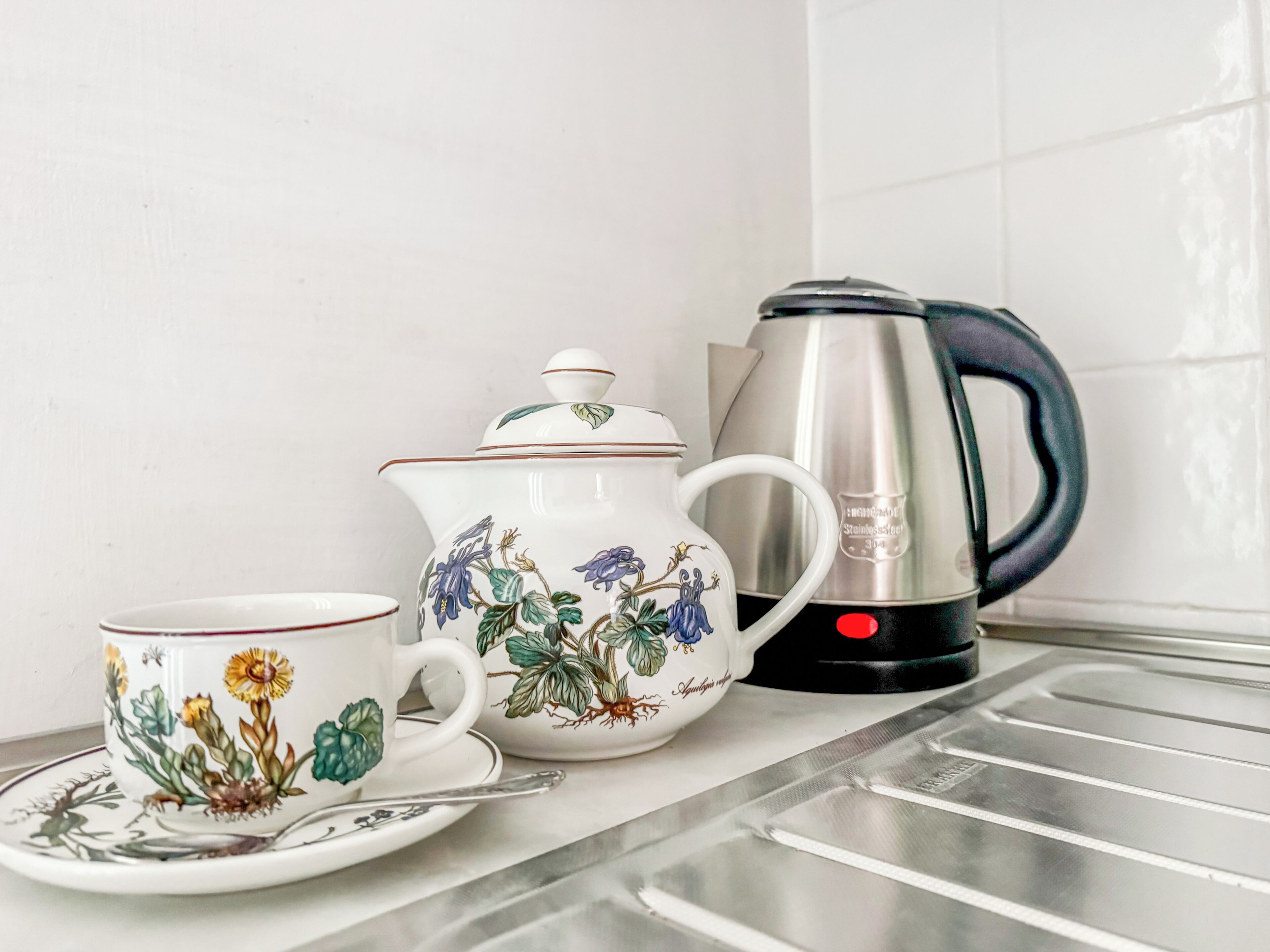 Una encimera de cocina con una tetera blanca, una taza a juego y un hervidor de acero inoxidable.