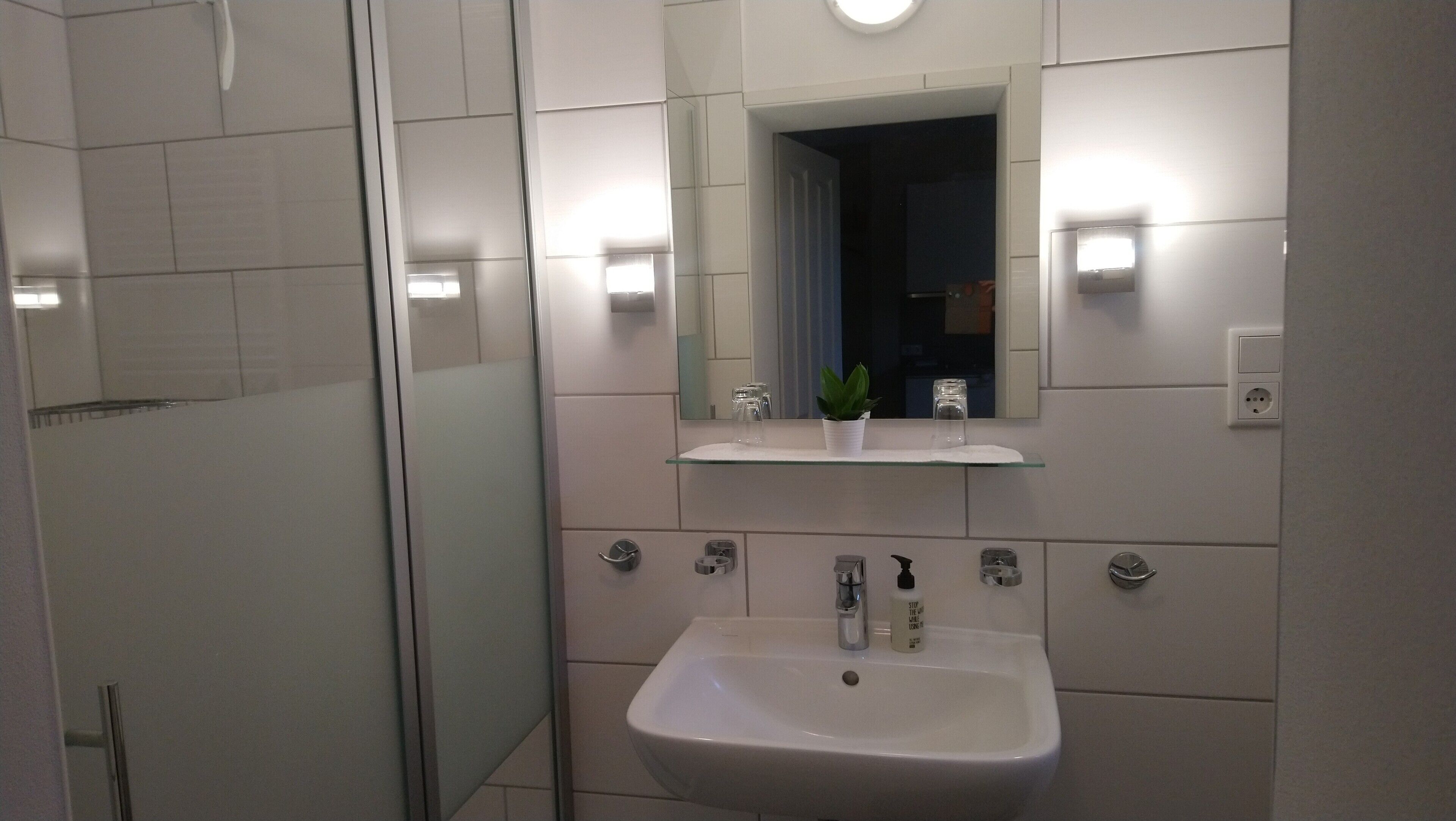 Uma casa de banho moderna com chuveiro em cabine de vidro, um lavatório branco com espelho em cima e uma pequena prateleira com uma planta.