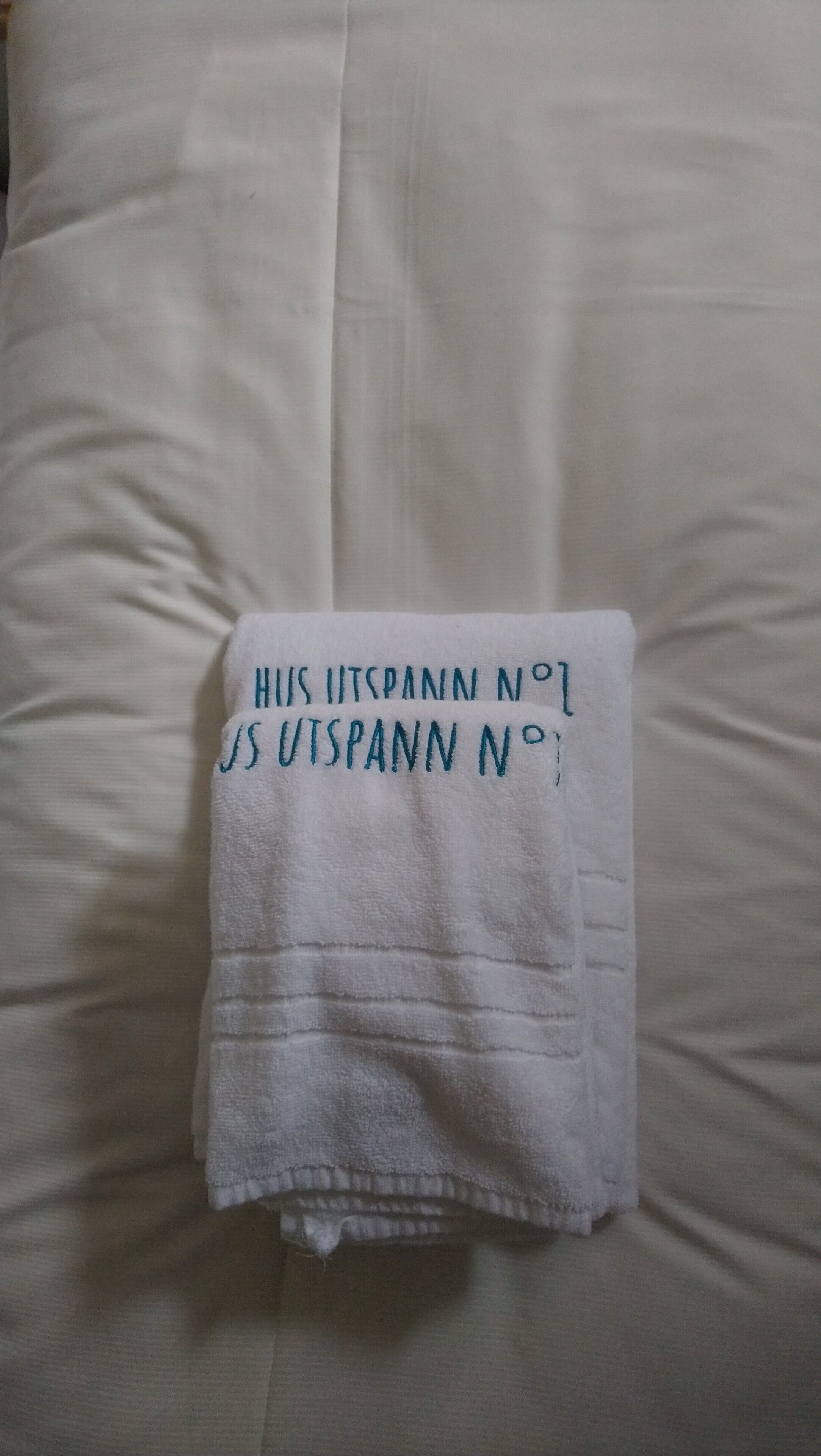 Ein gefaltetes weißes Handtuch mit Schrift auf einem Bett mit weißer Bettwäsche.
