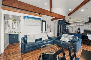 A modern living room with a blue sofa set, a glass coffee table, and a kitchen area with dark cabinetry and a black stove. - Lakefront Chalet in Boulder Bay! | Hot Tub | Fire Pit | Amazing Lake Views (Big Bear Lake)