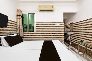 A hotel room with a bed, a window with curtains, an air conditioning unit, a mirror, and a small table with a lamp. - Deluxe Ayush Villa (Kolkata)