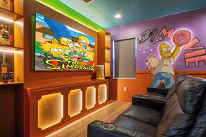 Ein Zimmer mit einer großen Wandmalerei mit den Simpsons, einem Flachbildfernseher, einem Ledersofa und einer Snackbar.