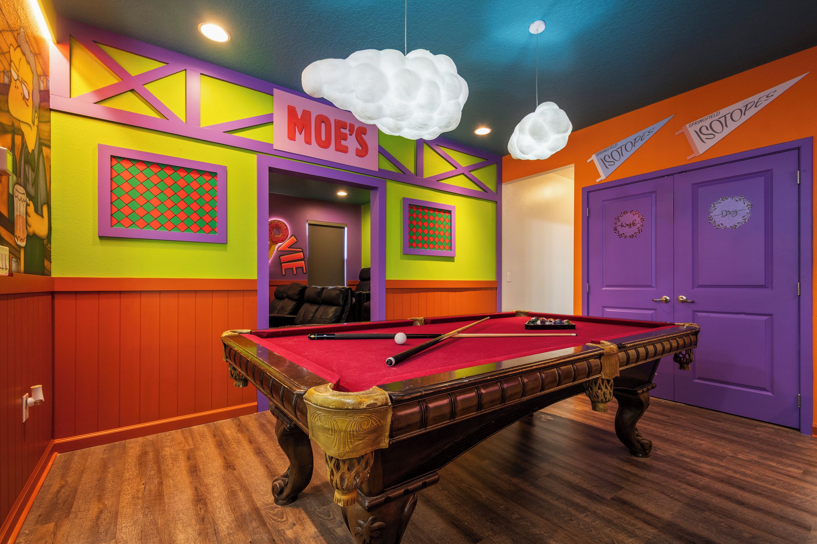 A pool table with a red felt surface, surrounded by walls with colorful geometric patterns and a purple door with a wreath.