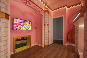 Ein pinkfarben gestaltetes Kinderzimmer mit Fernseher, der Zeichentrickfiguren zeigt, einem Spielbereich mit einer kleinen Modelleisenbahn und einer Tür zu einem anderen Raum.