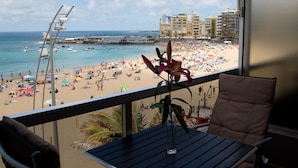 A balcony with a view of a crowded beach, a chair, and a table. - Vacation Studio 'Terraza A La Playa' with Wi-Fi (Las Palmas de Gran Canaria)