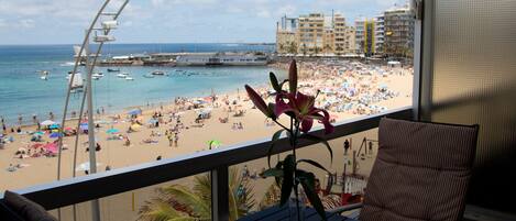 A balcony with a view of a crowded beach, a chair, and a table.