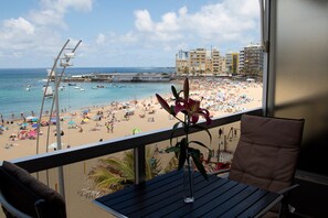 Ein Balkon mit Blick auf einen belebten Strand, ein Stuhl und ein Tisch.