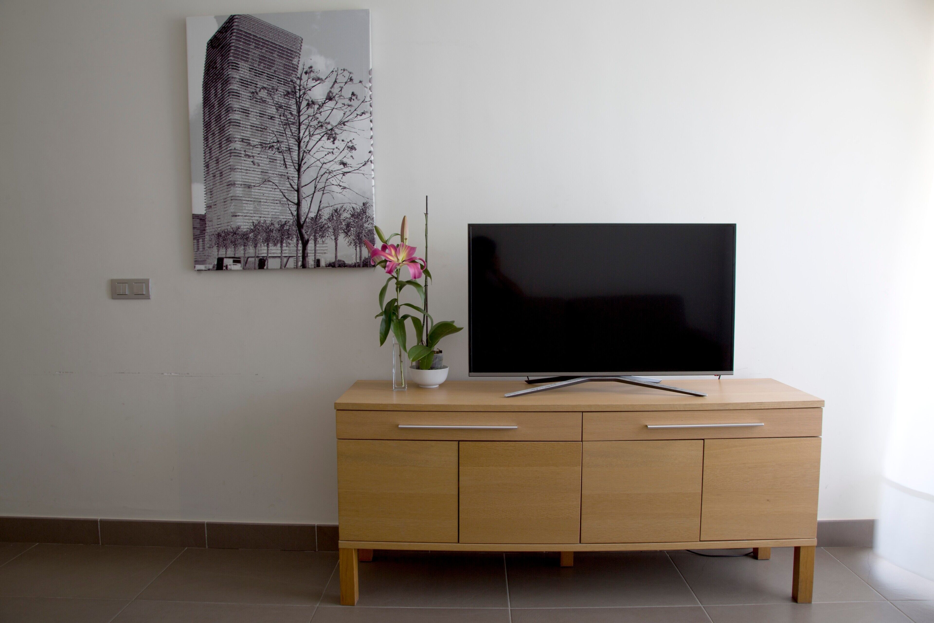 Uma sala de estar moderna com uma televisão de tela plana em um armário de madeira, um vaso com flores e uma pintura com vista para a cidade na parede.