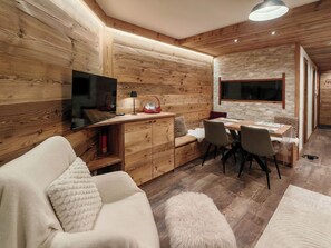 Ein gemütliches Zimmer mit Holzvertäfelung, einer Couch, einem Esstisch mit Stühlen und einem an der Wand befestigten Fernseher.