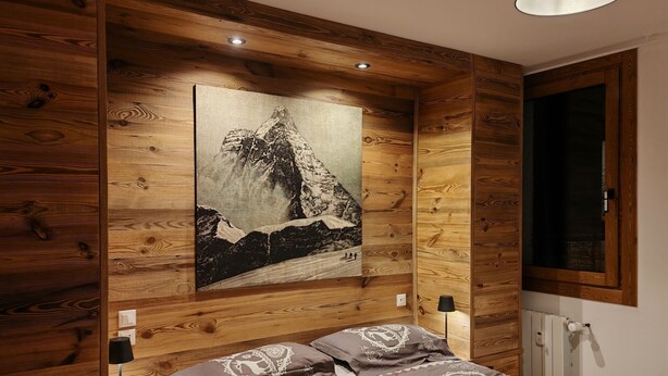 A bedroom with a wooden headboard, a painting of a mountain, a bed with patterned bedding, and a bedside lamp. - Apartment at the foot of the legendary Matterhorn in the Cervina–Zermatt area. (Breuil-Cervinia)