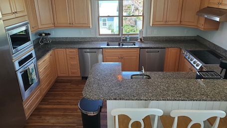 Una cocina con encimeras de granito, electrodomésticos de acero inoxidable, armarios de madera, un ventanal con vistas y un cubo de basura.