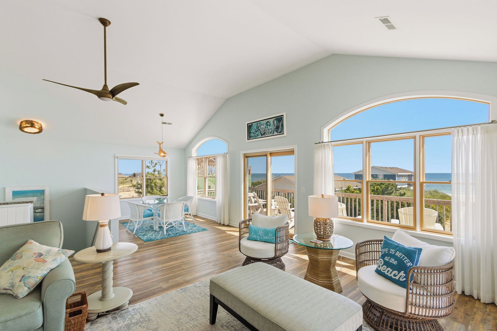 A spacious living room with a high ceiling, large windows offering ocean views, a dining area with a glass table, and a cozy seating arrangement.