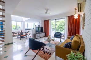 A modern living room with a sofa, a coffee table, and a television. There is a dining area with chairs and a table. - Modern 2bhk in Siolim With Pool Near Hotspots (Mapusa)