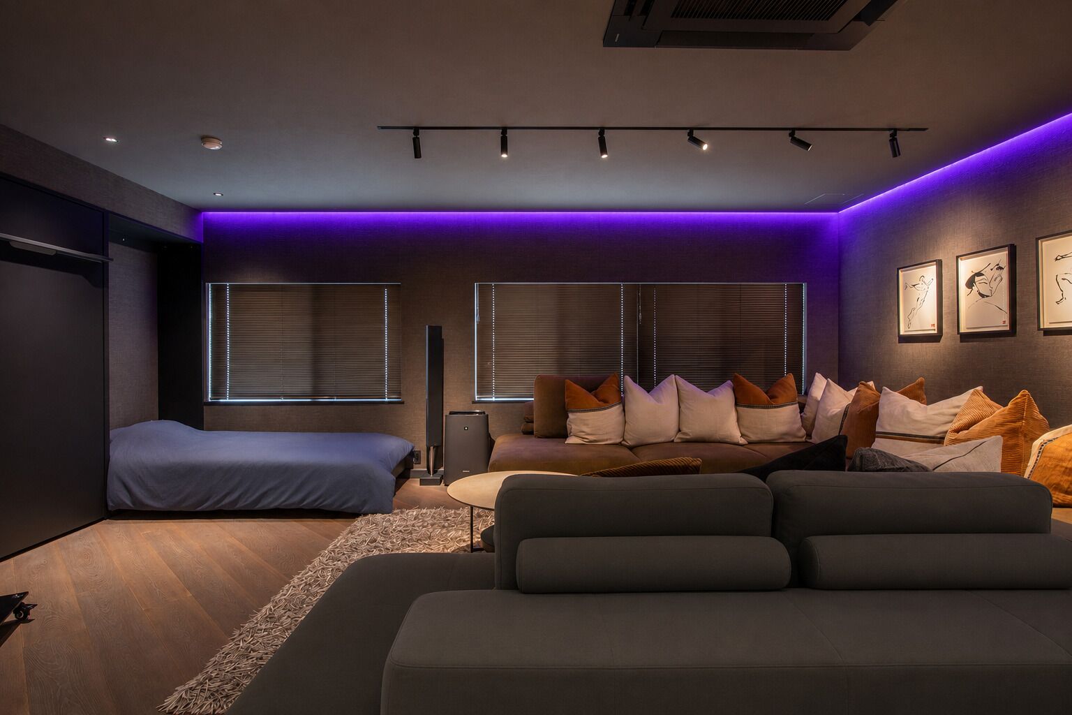 A modern bedroom with a bed, a sofa, and a desk. There are windows with blinds, a ceiling light, and a wall-mounted air conditioner.