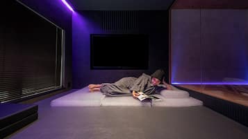 A person lying on a white couch in a room with a large flat-screen TV mounted on the wall, a window with blinds, and a purple ambient light strip.