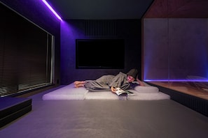 A person lying on a white couch in a room with a large flat-screen TV mounted on the wall, a window with blinds, and a purple ambient light strip. - Kanako Penthouse Sauna & Rooftop @ Monzen-Nakacho (Koto City)