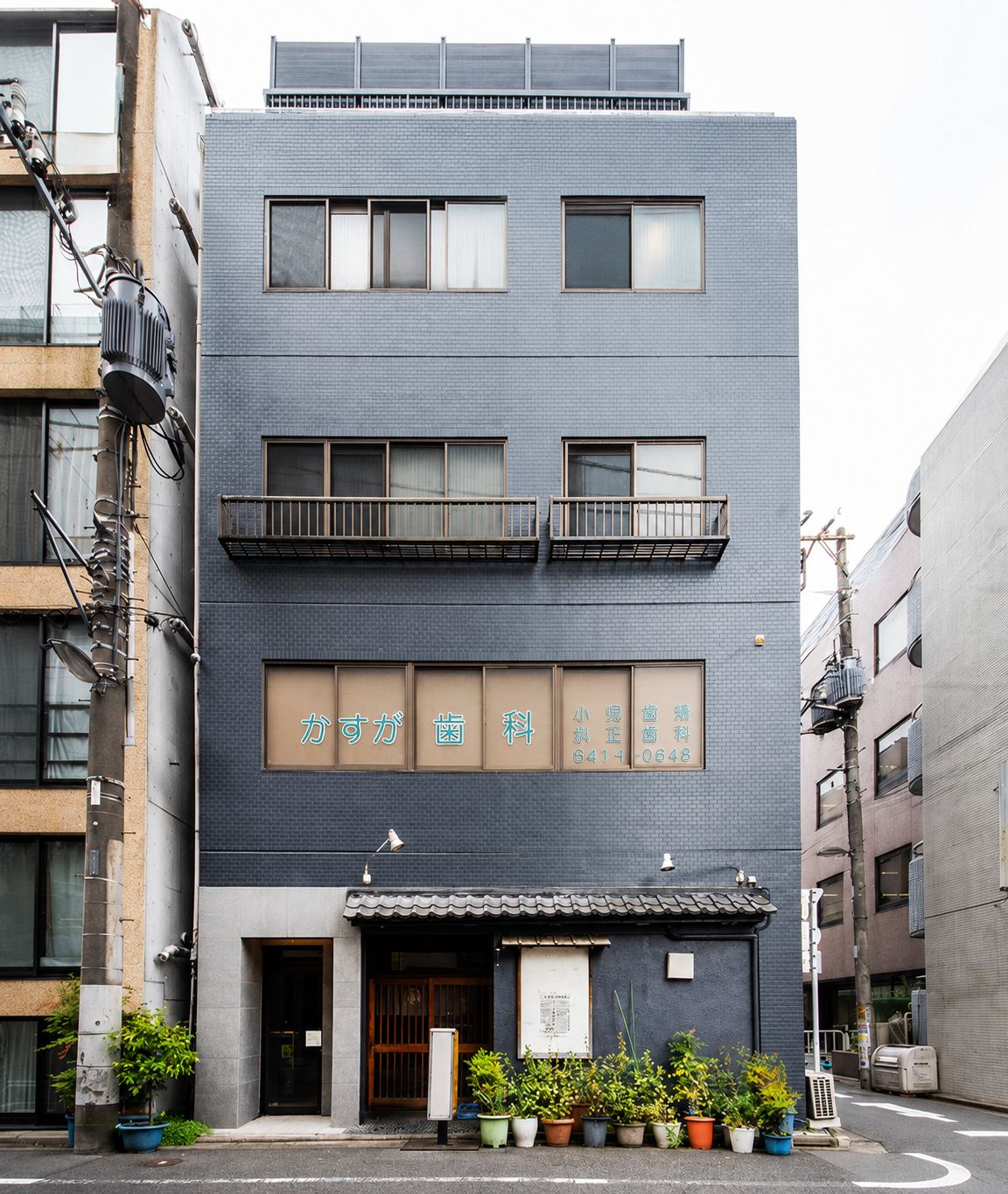 A multi-story building with a dark facade, multiple windows, and a ground-level entrance with potted plants.