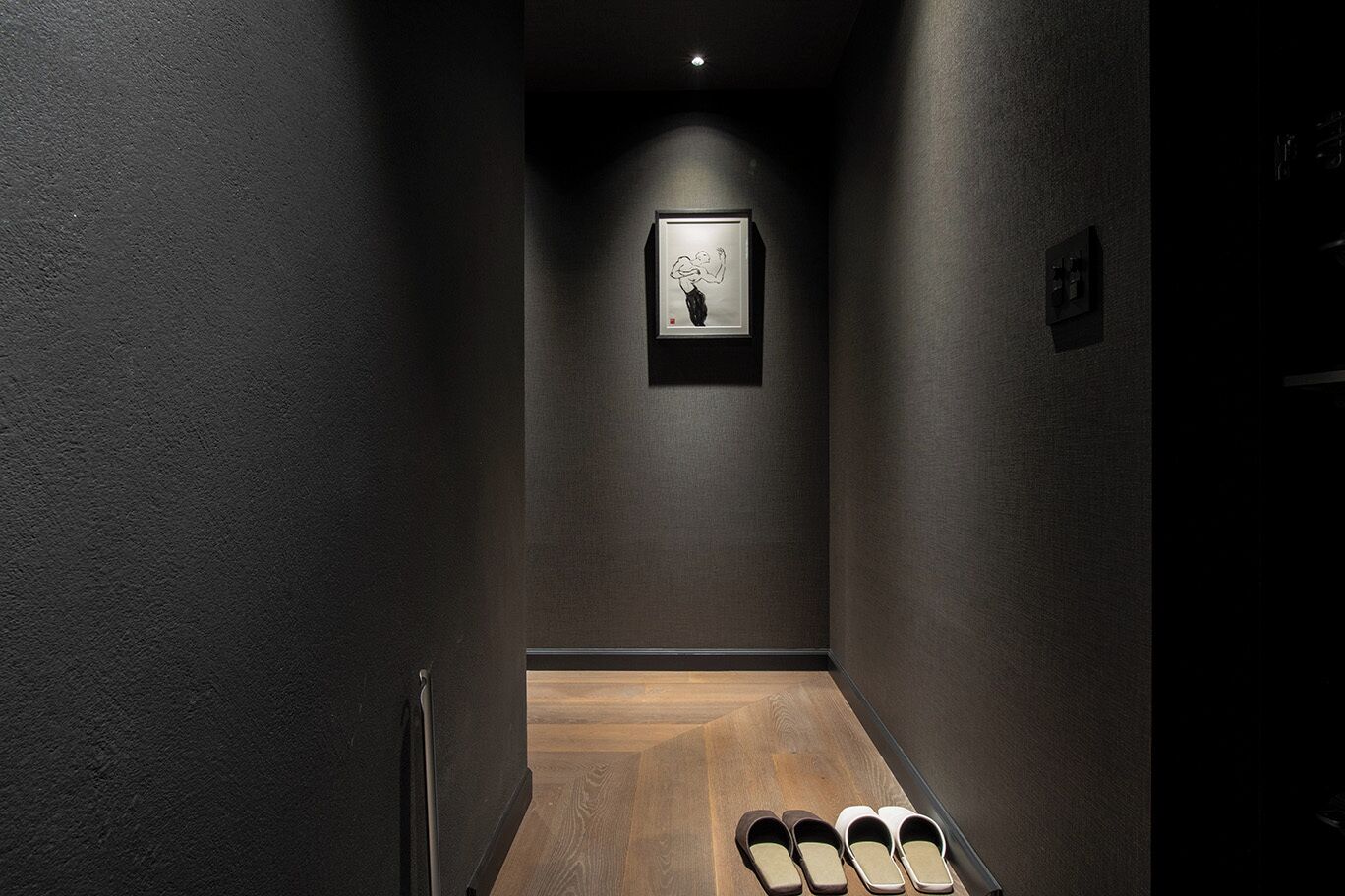 A narrow hallway with a wooden floor, a framed artwork on the wall, and a pair of slippers at the end.