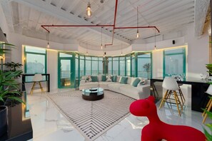Uma sala de estar moderna com um sofá branco, uma mesa de centro de vidro e uma poltrona vermelha. Há grandes janelas com vista para a cidade.