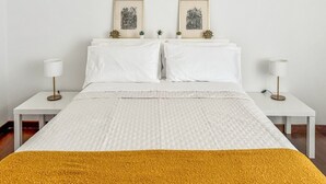 Un llit de matrimoni ben fet amb un edredó blanc i una manta de color mostassa, flanquejat per dues taules de nit amb làmpades, i una obra d'art a la paret sobre el llit.