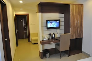 A hotel room with a TV mounted on the wall, a desk with a chair, a small trash bin, and a door leading to another room. - NIRVANA HOTEL (Varanasi)