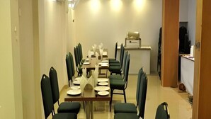 Un espace repas avec une longue table dressée pour plusieurs personnes, des chaises disposées autour, et un petit coin cuisine à l’arrière-plan.