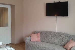 Een woonkamer met een grijze bank, twee kussens, een flatscreen televisie aan de muur en een deur met gordijn.