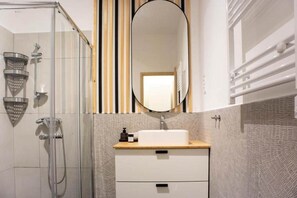 A bathroom with a glass shower enclosure, a white sink with a wooden countertop, and a large mirror above the sink. - MidTerm - Rózsa Apartments - Jeremy B (Budapest)