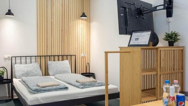 Quarto moderno com uma cama, TV montada e uma prateleira de madeira com garrafas.