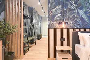 A modern room with a bed, a small table, a bench, and a wall with a tropical leaf pattern. - IMCT - Urban Jungle Apartments 01 (Budapest)