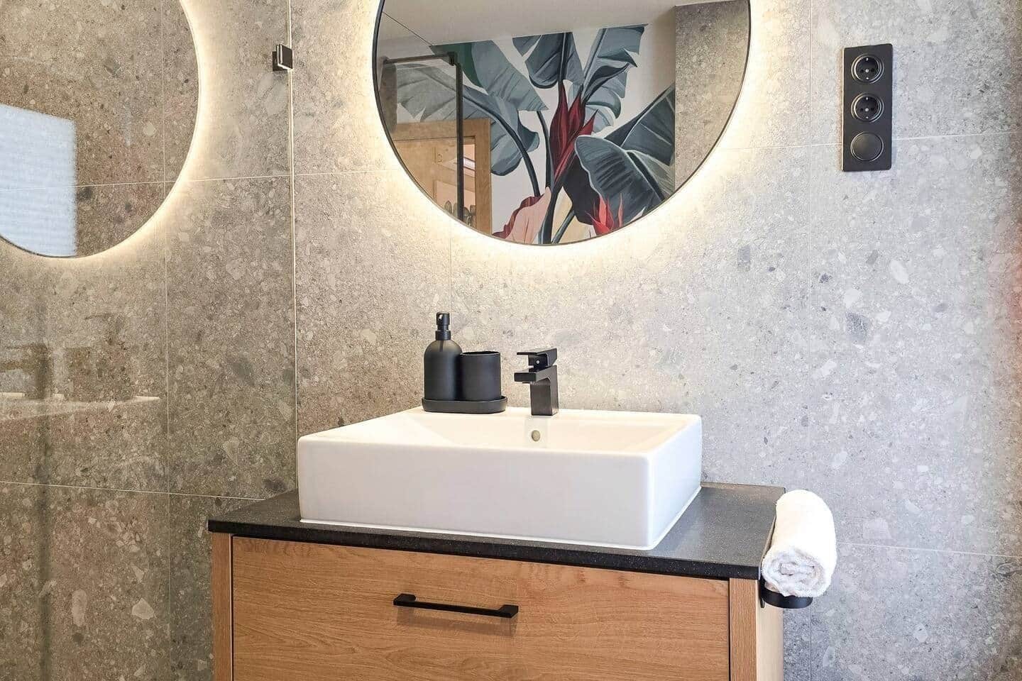 A bathroom with a white sink, black faucet, and a round mirror with a plant reflection.