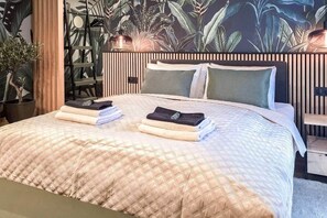 A hotel room with a bed, a nightstand, a potted plant, and a wall with a tropical leaf pattern. - IMCT - Urban Jungle Apartments 01 (Budapest)